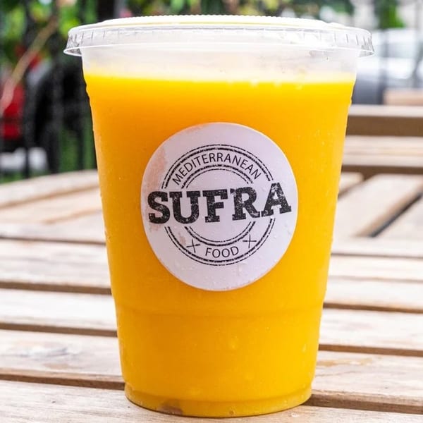 A plastic cup of bright orange juice with a "Sufra Mediterranean Food" sticker sits on a wooden table.